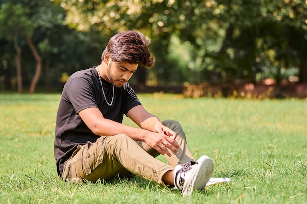 Porträt eines attraktiven jungen indischen Mannes in schwarzem T-Shirt und silberner Halskette, der auf grünem Rasen sitzt