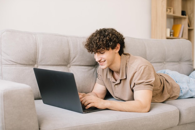 Porträt eines attraktiven jungen Mannes mit lockigem Haar, der auf dem Sofa liegt und Laptop verwendet