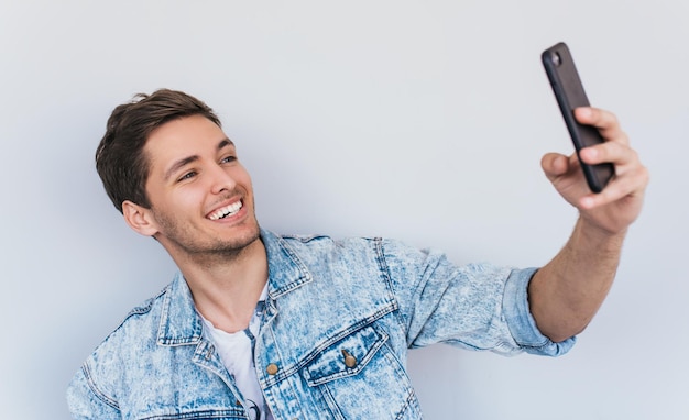 Porträt eines attraktiven kaukasischen Mannes in stilvoller Pose für Selfie mit glücklichem Lächeln in Jeansjacke vor weißem Studiowandhintergrund Lebensstil der Menschen und modernes Technologiekonzept