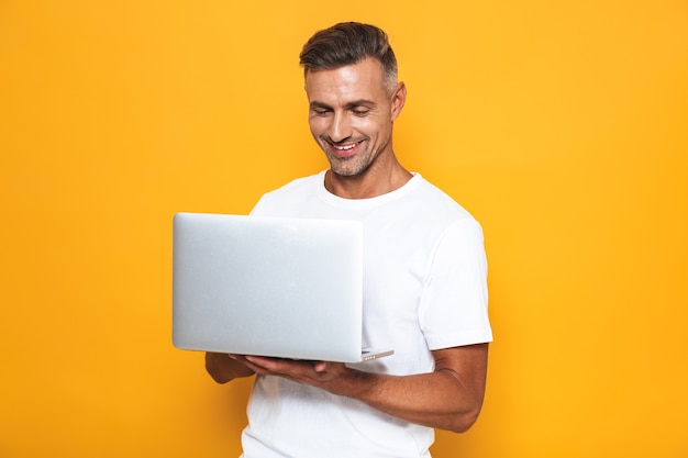 Porträt eines attraktiven Kerls der 30er Jahre in weißem T-Shirt, der einen silbernen Laptop hält und verwendet, isoliert auf Gelb