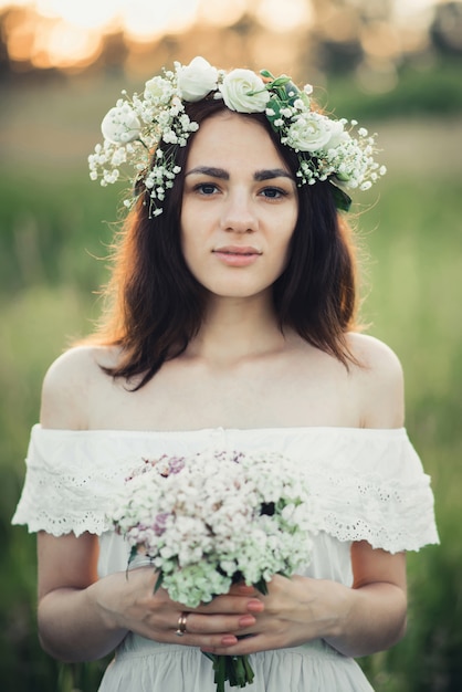 Porträt eines attraktiven Mädchens im weißen Kleid mit einem Blumenstrauß und einem Kranz im Sommer