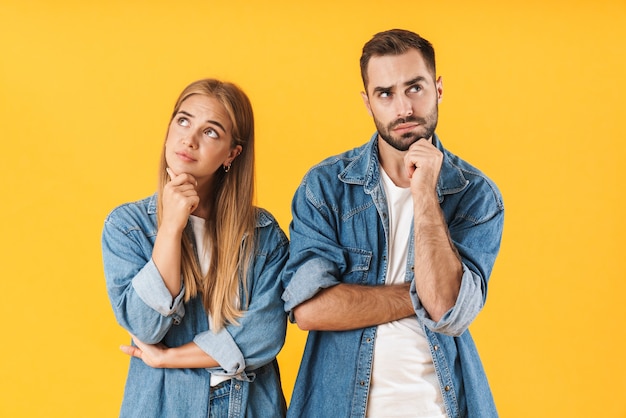 Porträt eines attraktiven nachdenklichen jungen Paares in Freizeitkleidung, das isoliert über gelber Wand steht und in beide Richtungen schaut
