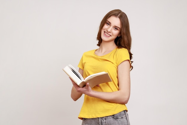 Porträt eines attraktiven Teenager-Mädchens, das ein Buch hält und lächelt und das Lesen genießt, einen Zeitplan im Notizblock erstellt, der mit glücklichem Ausdruck in die Kamera blickt Studioaufnahme isoliert auf grauem Hintergrund