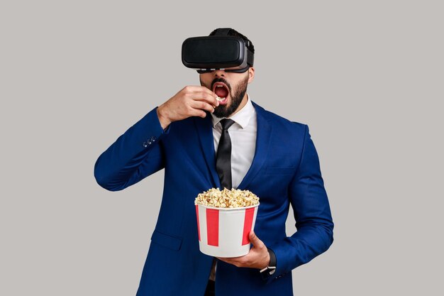 Porträt eines aufgeregten, erstaunten bärtigen Geschäftsmannes mit VR-Headset, der einen Film mit Popcorn sieht, der einen leckeren Snack im Anzug im offiziellen Stil isst Studioaufnahme isoliert auf grauem Hintergrund