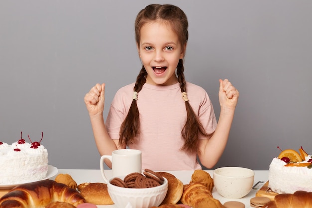 Porträt eines aufgeregten, fröhlichen, überglücklichen kleinen Mädchens mit Zöpfen, das isoliert über grauem Hintergrund am Tisch sitzt, geballte Fäuste, die bereit sind, viele Süßigkeiten und hausgemachtes Gebäck zu essen