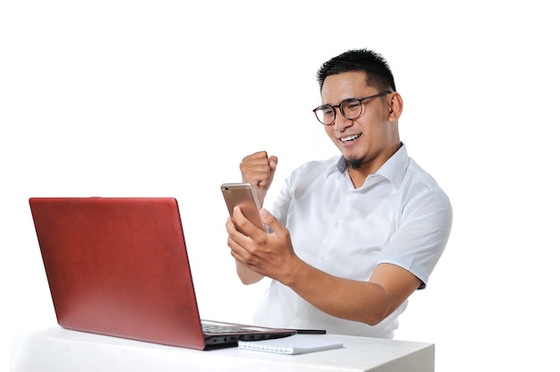 Porträt eines aufgeregten jungen asiatischen Mannes, der in der Nähe des Laptops auf dem Schreibtisch sitzt, hat die Arbeit als Online-Freiberufler erfolgreich abgeschlossen und die Zahlung erhalten, isoliert auf weiß