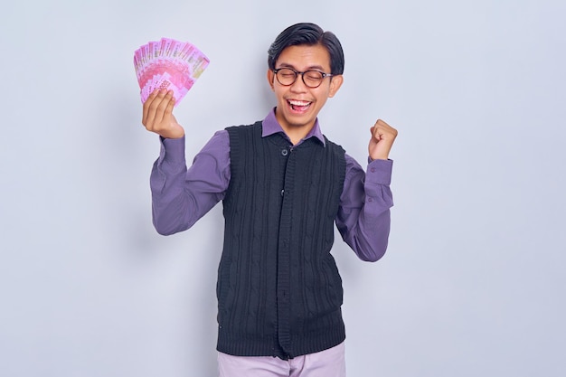 Porträt eines aufgeregten jungen, gutaussehenden asiatischen Geschäftsmannes, der Bargeld in Rupiah-Banknoten hält, die auf weißem Hintergrund isoliert sind Menschen-Lifestyle-Konzept