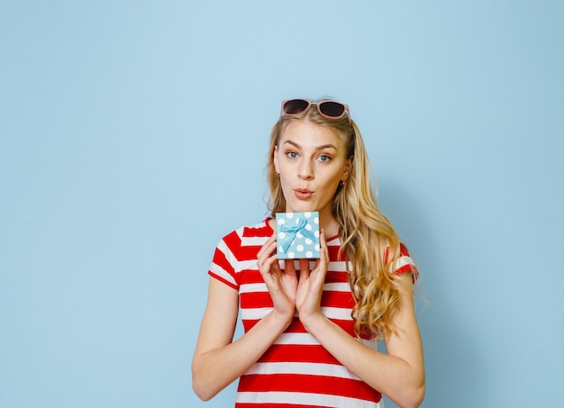 Porträt eines aufgeregten jungen schönen blonden Mädchens, das ein Geschenk hält und über blauem Hintergrund glücklich ist