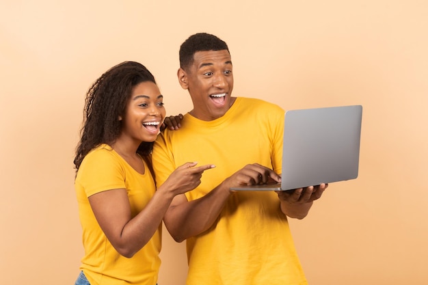 Porträt eines aufgeregten schwarzen Paares, das zusammen auf einen Laptop zeigt und Gadget mit aufgeregtem pfirsichfarbenem Hintergrund betrachtet