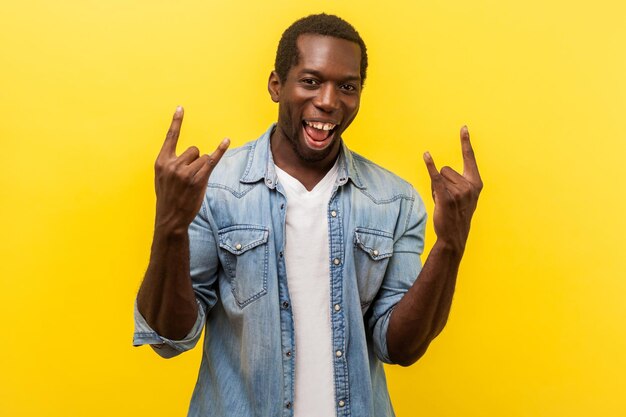 Porträt eines aufgeregten, verrückten, fröhlichen Mannes in Jeans-Freizeithemd mit aufgerollten Ärmeln, der Rock'n'Roll-Geste mit erhobenen Händen zeigt, die in die Kamera lächeln, Studioaufnahme einzeln auf gelbem Hintergrund