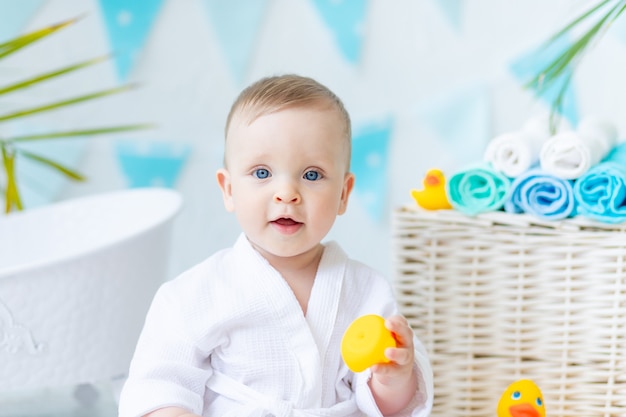 Porträt eines Babys, das nach dem Baden in der Badewanne in einem weißen Gewand mit einer Gummiente und einem Handtuch sitzt, das Konzept der Hygiene und des Waschens.