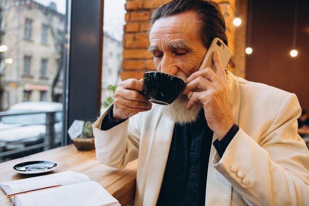 Porträt eines bärtigen älteren Mannes, der Kaffee trinkt und Smartphone im Café benutzt. Porträt des glücklichen grauen bärtigen Mannes, der im Café sitzt.