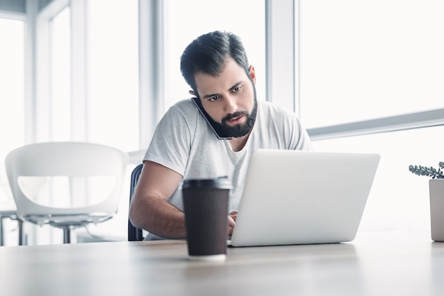 Porträt eines bärtigen dunkelhaarigen Mannes, der in einem weißen Büro sitzt und beim Telefonieren auf seinen Laptop-Bildschirm schaut. Seine Tasse Kaffee steht vor ihm