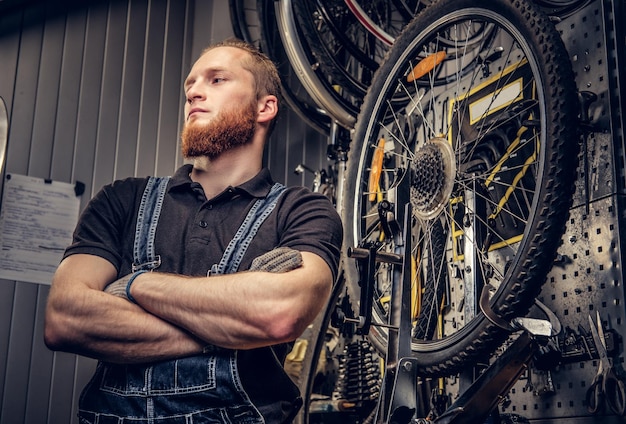 Porträt eines bärtigen Fahrradmechanikers mit rotem Kopf in einer Werkstatt mit Fahrradteilen und Rad auf einem Hintergrund.
