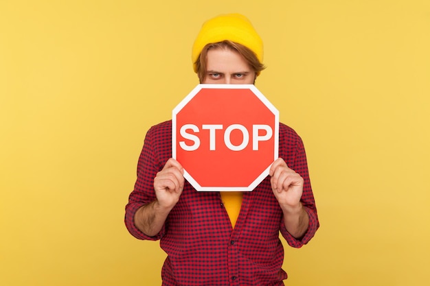 Foto porträt eines bärtigen hipster-typen mit mütze und kariertem hemd, der die hälfte des gesichts mit rotem verkehrsschild bedeckt, warnung zu gehen, verbotskonzept. studioaufnahme im innenbereich isoliert auf gelbem hintergrund.