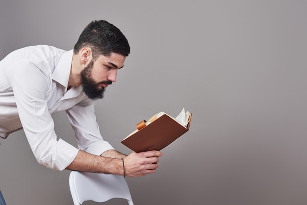 Porträt eines bärtigen jungen Mannes, der ein weißes Hemd trägt und einen offenen Planer und einen Stift hält. Eine graue Wand