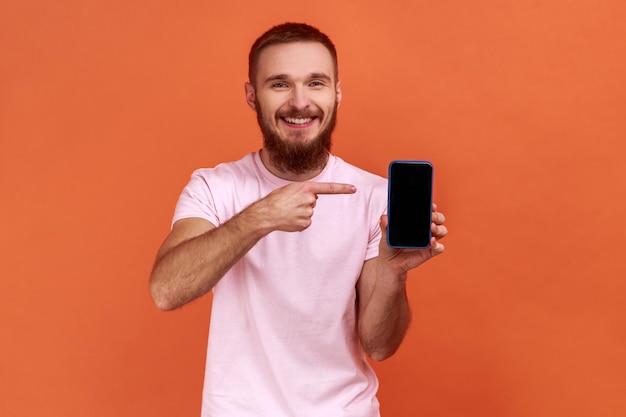 Porträt eines bärtigen Mannes, der auf ein Handy zeigt und in die Kamera lächelt, ein Gadget oder eine mobile Anwendung empfiehlt und ein rosa T-Shirt trägt. Studioaufnahme im Innenbereich isoliert auf orangefarbenem Hintergrund.