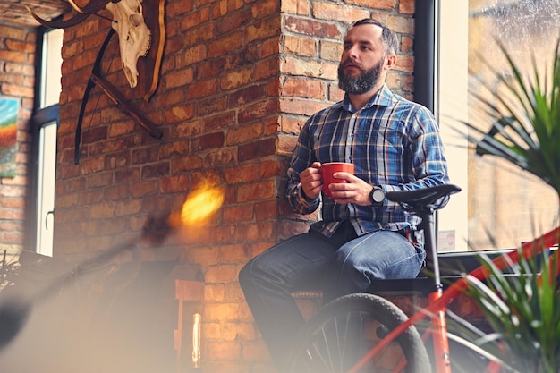 Porträt eines bärtigen Mannes in einem blauen Fleecehemd, trinkt heißen Kaffee in der Nähe des Fensters im Ruheraum mit Loft-Interieur.