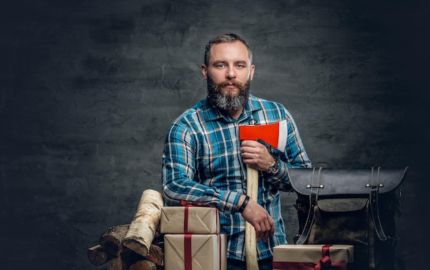 Porträt eines bärtigen Mannes mittleren Alters hält eine Axt und steht neben einem Tisch mit Weihnachtsgeschenkboxen und Brennholz auf grauem Hintergrund.