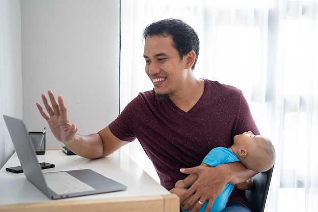 Porträt eines beschäftigten Vaters, der von zu Hause aus arbeitet, während er sein Baby hält