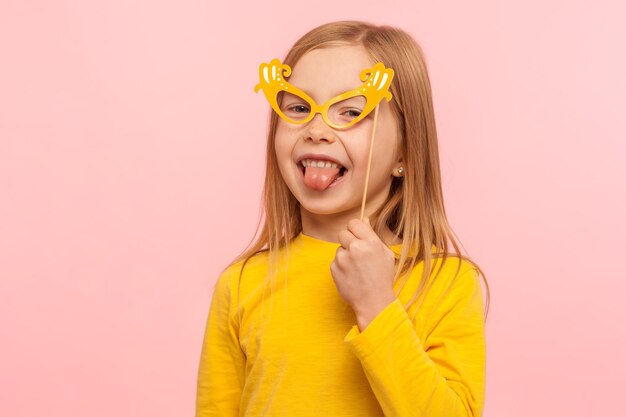 Porträt eines bezaubernden kleinen Mädchens aus Ingwer, das die Augen mit einer Papierbrille bedeckt und die Zunge herausstreckt, ein ungezogenes Kind, das Spaß hat und eine Maskerademaske trägt. Studioaufnahme im Innenbereich isoliert auf rosa Hintergrund