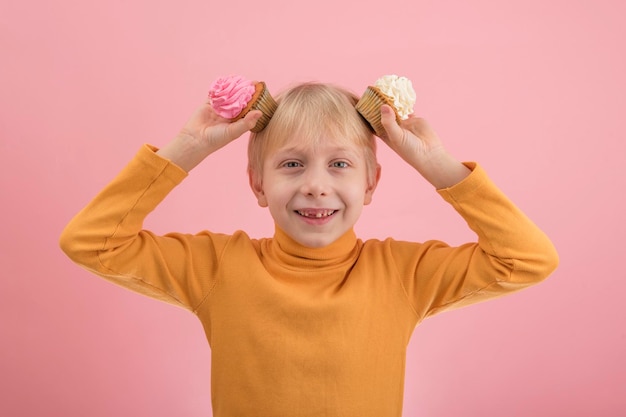 Porträt eines blonden Jungen in heller Kleidung mit Cupcakes in den Händen auf rosa Hintergrund. Kinderfest mit Süßigkeiten.