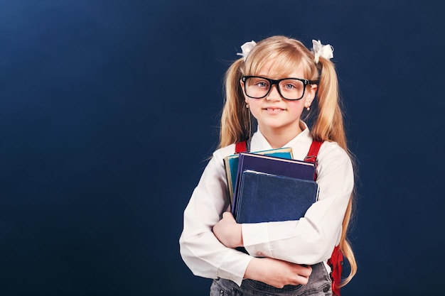 Porträt eines blonden, lächelnden Schulmädchens mit Brille und Rucksack, das Bücher vor blauem Hintergrund hält