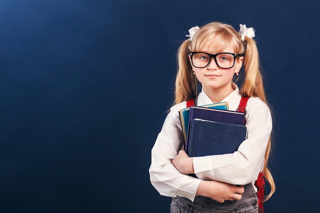 Porträt eines blonden Schulmädchens trägt eine Brille und einen Rucksack, der Bücher vor blauem Hintergrund hält