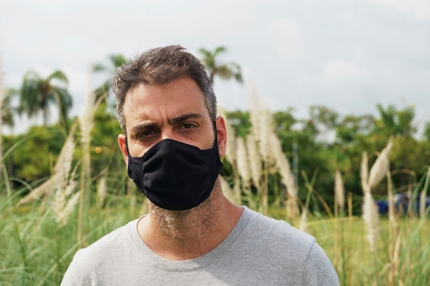 Porträt eines brasilianischen Mannes, der eine Schutzmaske trägt, während er im Park steht. Porträt eines mittleren erwachsenen Mannes im Freien, der eine schwarze Maske trägt, um sich vor einer Coronavirus-Pandemie zu schützen. Natur im Hintergrund.