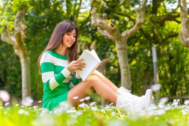 Porträt eines brünetten kaukasischen Mädchens, das ein Buch in der Natur liest und auf dem Gras sitzt und einen grün-weißen Pullover trägt