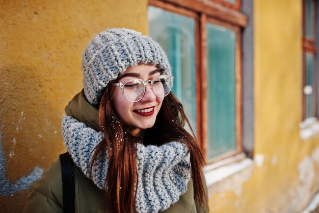 Porträt eines brünetten Mädchens in grauem Schal und Hut, Brille bei kaltem Wetter mit Sonnenschein gegen orangefarbene Wand des alten Hauses.