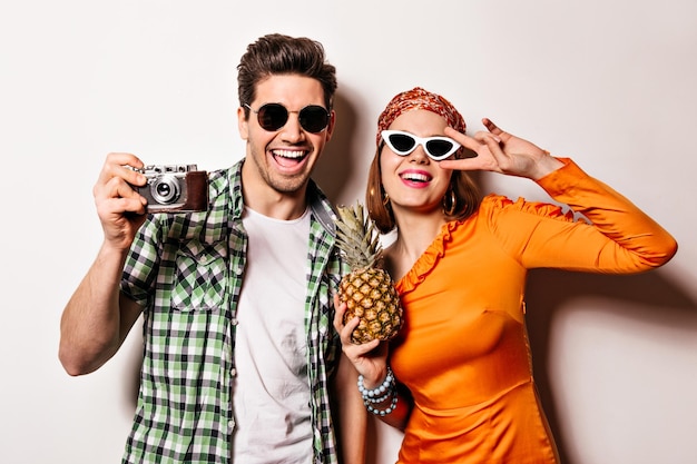 Porträt eines Brunet-Mannes, der mit einer Retro-Kamera fotografiert, und einer Frau in orangefarbenem Kleid und Stirnband Mädchen mit Brille hält Ananas und zeigt Friedenszeichen