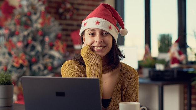 Porträt eines Büroangestellten mit Weihnachtsmütze während der Winterferien, der am Schreibtisch am Laptop arbeitet, bevor er Heiligabend feiert. Frau, die sich festlich im Raum fühlt, der mit Weihnachtsornamenten geschmückt ist.