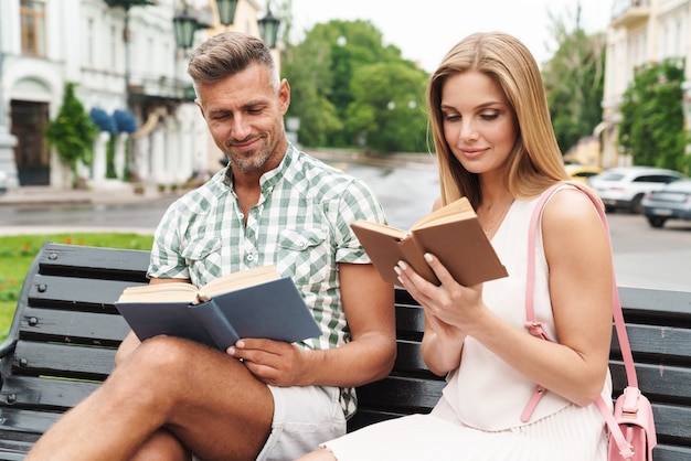 Porträt eines charmanten jungen Paares in Sommerkleidung, das zusammen lächelt und Bücher liest, während es auf der Bank in der Stadtstraße sitzt