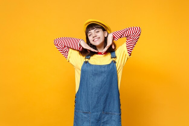 Porträt eines charmanten Mädchen-Teenagers in französischer Baskenmütze, Denim-Sommerkleid, das die Hände in der Nähe des Gesichts isoliert auf gelbem Wandhintergrund im Studio hält. Menschen aufrichtige Emotionen, Lifestyle-Konzept. Mock-up-Kopienbereich.