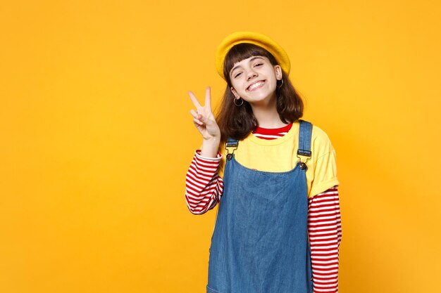 Porträt eines charmanten Mädchen-Teenagers in französischer Baskenmütze und Denim-Sommerkleid mit Siegeszeichen isoliert auf gelbem Wandhintergrund im Studio. Menschen aufrichtige Emotionen, Lifestyle-Konzept. Mock-up-Kopienbereich.