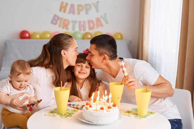 Porträt eines charmanten Mädchens, das Geburtstag mit ihrer Familie feiert, ein Kind, das zu Hause Glückwünsche erhält, Eltern, die ihre Tochter küssen und mit ihrem Tag gratulieren.