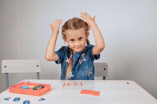 Porträt eines charmanten niedlichen und fröhlichen Mädchens, das an einem weißen Tisch sitzt, umgeben von Rätseln und ihre Hände in der Erhebung hebt. Kindheitskonzept. Foto mit Lärm