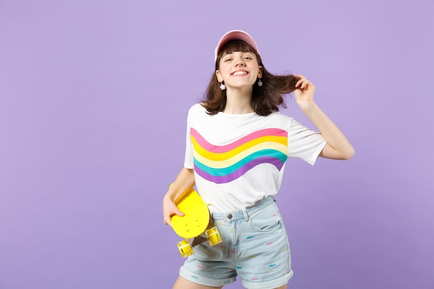 Porträt eines charmanten Teenager-Mädchens in lebhafter Kleidung mit gelbem Skateboard, das Haare isoliert auf violettem Pastellwandhintergrund im Studio hält. Menschen aufrichtige Emotionen, Lifestyle-Konzept. Mock-up-Kopienbereich.