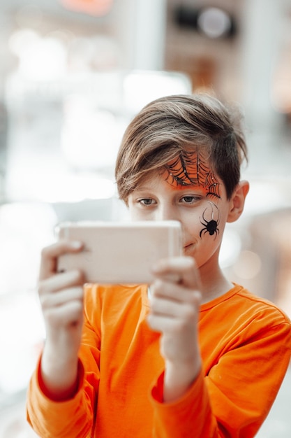 Porträt eines ehrlichen, authentischen Zoomer-Jungen mit Schminken mit dem Handy auf der Halloween-Party