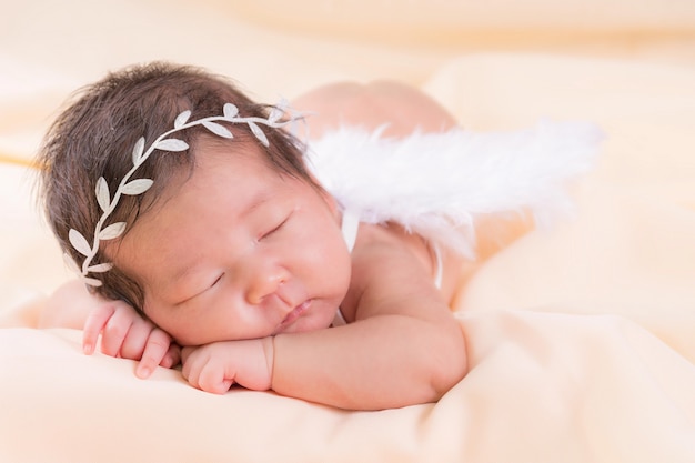 Porträt eines einen Monat alten schlafenden, neugeborenen Mädchens. Sie trägt ein weißes Kronenstirnband, Engelsflügel und schläft auf einer cremefarbenen Decke. Konzept Portraitstudio Mode Neugeborenes.