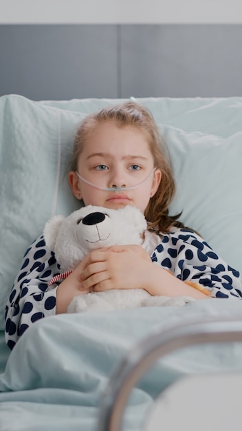 Porträt eines einsamen kranken kleinen Kindes, das im Bett ruht und einen Teddybären in den Händen hält und während der Genesungsuntersuchung in der Krankenstation in die Kamera schaut. Kind im Krankenhaus, das auf medizinische Behandlung wartet