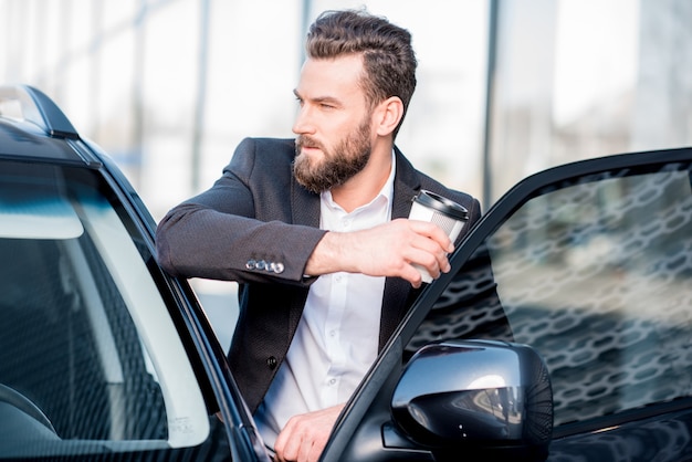 Porträt eines eleganten Geschäftsmannes, der mit Kaffeetasse nahe dem Auto steht