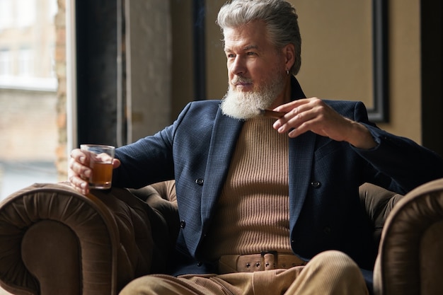 Porträt eines eleganten reifen Geschäftsmannes mit Bart, der beiseite schaut, während er ein Glas Whisky hält und