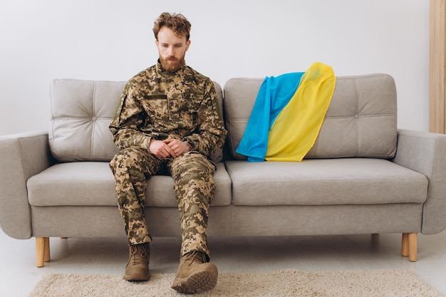 Porträt eines emotionalen jungen ukrainischen Patriotensoldaten in Militäruniform, der im Büro auf einem Sofa mit gelber und blauer Flagge sitzt