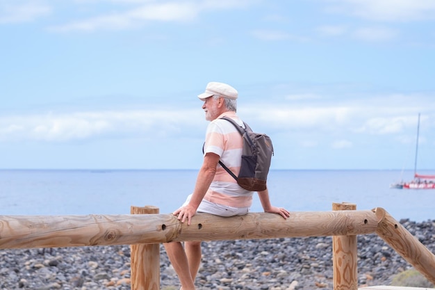 Porträt eines entspannten kaukasischen bärtigen Mannes mit Hut, der am Strand sitzt und den Horizont betrachtet, während er Urlaub oder Ruhestand genießt Horizont über dem Wasserkopierraum