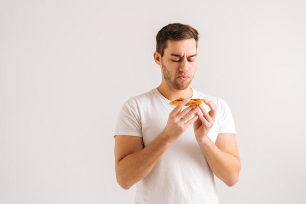 Porträt eines enttäuschten verärgerten jungen Mannes, der ein Stück Pizza mit schrecklichem Geruch hält, verdorbenes, unappetitliches Essen riecht, unzufrieden mit der Lebensmittelqualität auf weißem, isoliertem Hintergrund im Studio.