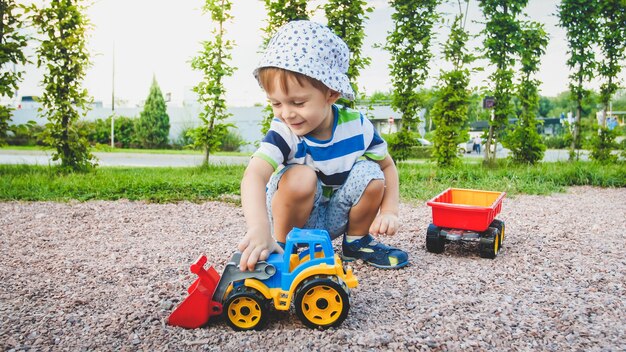 Porträt eines entzückenden 3-jährigen kleinkindjungen, der mit spielzeug-lkw mit anhänger auf dem spielplatz im park spielt