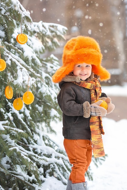 Porträt eines entzückenden kleinen Jungen in einem orangefarbenen Hut, der im Winter eine Orange in die Hand nimmt Orangenfrüchte wachsen auf dem Weihnachtsbaum