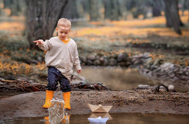 Porträt eines entzückenden kleinen Jungen in Gummistiefeln startet Papierboote in einer Pfütze im Frühling Origami